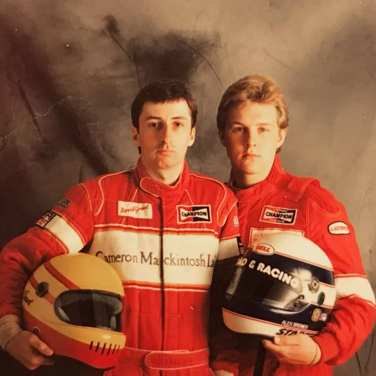 Alex, right, and team-mate David Grant in red Cameron Mackintosh Ltd race suits, each holding a helmet; Alex's helmet carries his name