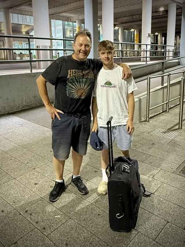Alex, smiling in a Here Comes The Sun T-shirt, with his arm around his son George, who stands beside a wheeled suitcase