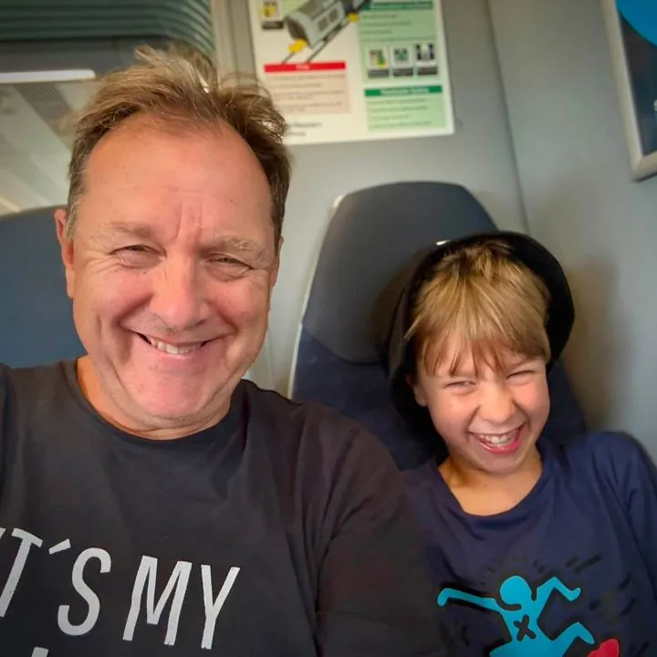 Selfie of a grinning Alex and his son Teddy, laughing in a cap, sitting side by side on a train
