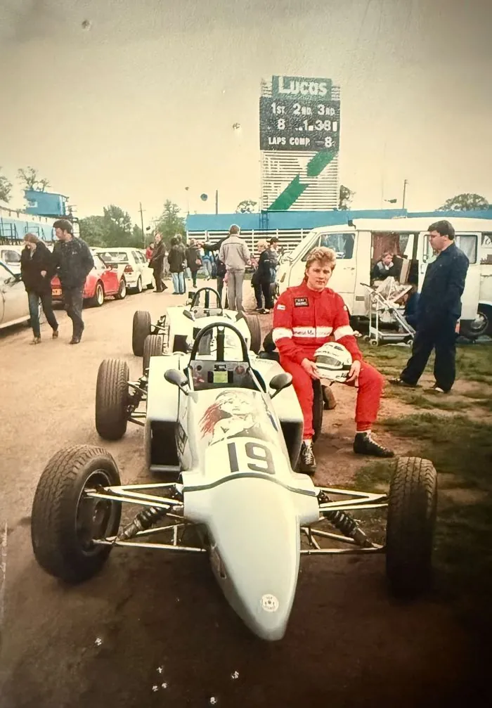 Alex in red race overalls, helmet in hand, sitting beside his white single-seater, number 19, in the paddock at Oulton Park
