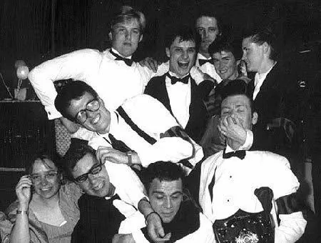 A crowd of young people in black tie and white shirts piled together, laughing, at a party