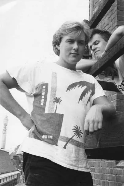 Alex in a palm-tree print T-shirt on a rooftop fire escape, a friend leaning on the rail behind him, the BT Tower in the distance