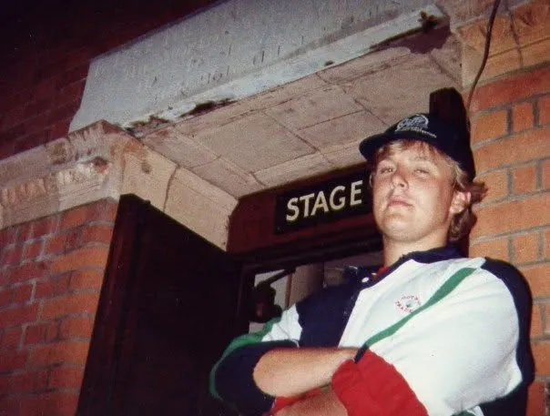 Alex in a cap and sports jacket, arms folded, beneath a sign reading STAGE over a brick doorway