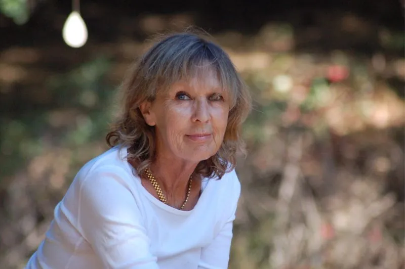 Alex's mother, Sylvia Schwadron, smiling in a white top, outdoors against a soft green and gold background