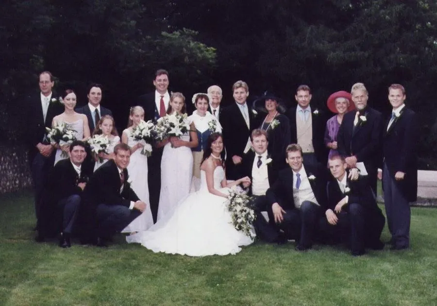 The wedding party on a lawn: Miriam in a white gown seated at the centre beside Alex, surrounded by bridesmaids, ushers and family