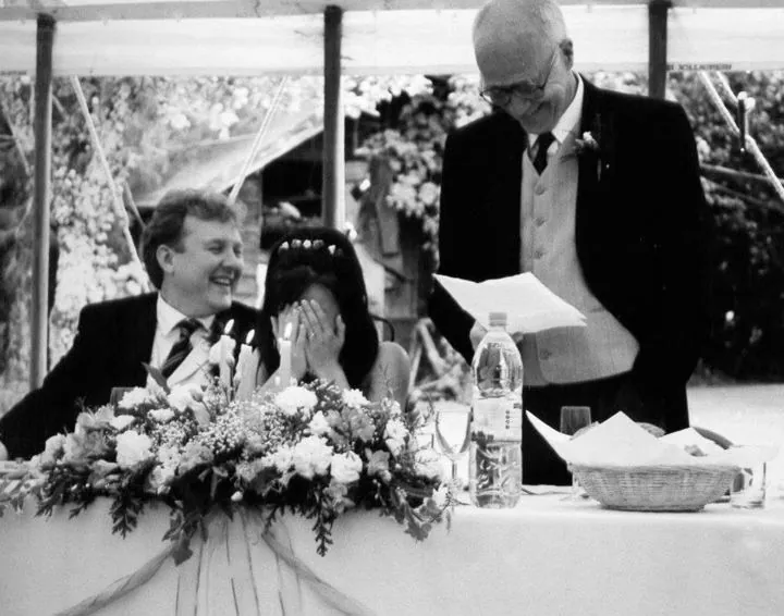 Black and white photo of Alex laughing and Miriam hiding her face in her hands as a guest gives a speech at the top table